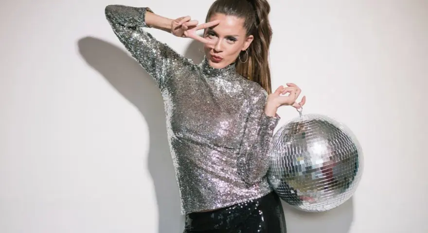 Kobieta w cekinowym topie i spodniach, z kulą dyskotekową w ręku, pozuje w stylu disco.