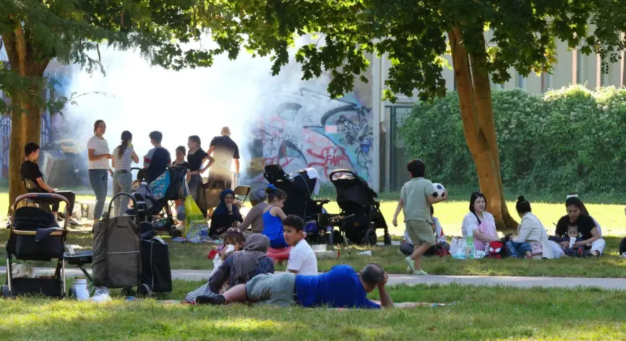 Familien genießen den Sommer auf einem Grillplatz in Berlin. Rauch steigt von einem Grill auf, während Kinder spielen und sich entspannen.