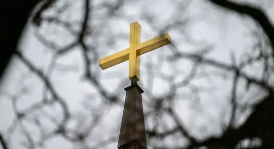 Goldenes Kreuz auf Kirchturmspitze, umrahmt von Ästen. Gedanken an die Kirchensteuer reduzieren.
