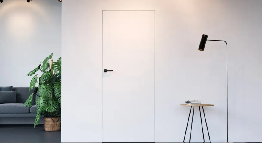Białe drzwi ukryte w ścianie z czarną klamką, obok stojąca lampa i stolik.