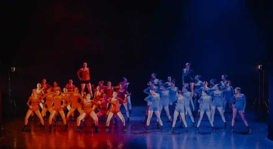 Grupa tancerzy na scenie, podzielona na dwie grupy: jedna w pomarańczowych strojach, druga w niebieskich. Nauka tańca nowoczesnego w pełnym blasku.