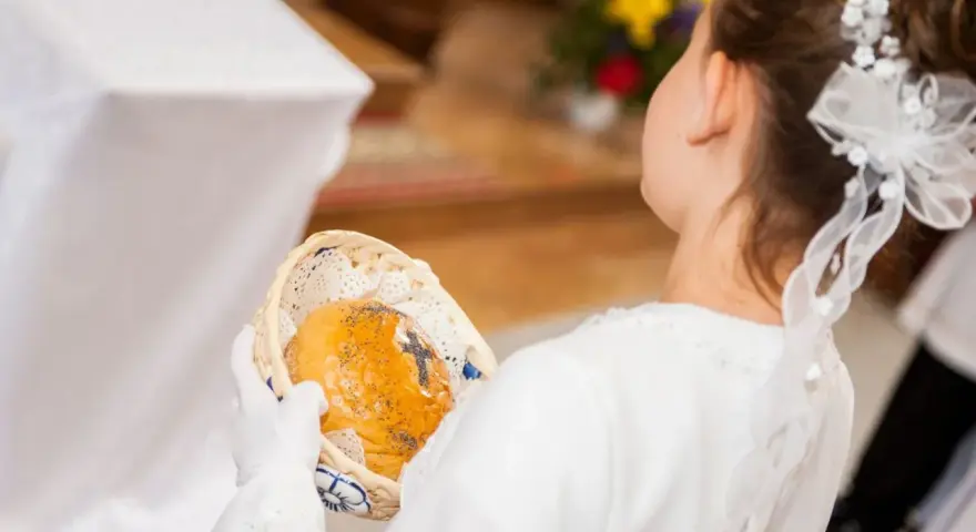 Dziewczynka w bieli trzyma koszyczek z opłatkiem. Zastanawiasz się, ile daje chrzestny na komunię?