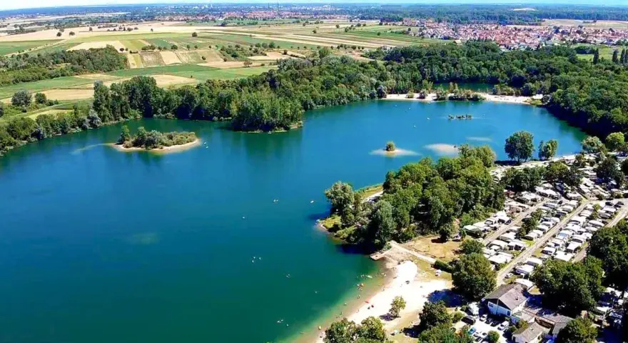 Der Mechtersheim See mit seiner türkisen Wasserfarbe, umgeben von grünen Bäumen und einem Campingplatz. Im Hintergrund erstrecken sich Felder und eine Stadt.