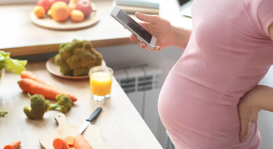 Ciężarna kobieta zmagająca się z brakiem apetytu w ciąży, szuka w telefonie przepisu na zdrowe jedzenie. Na blacie warzywa i sok.