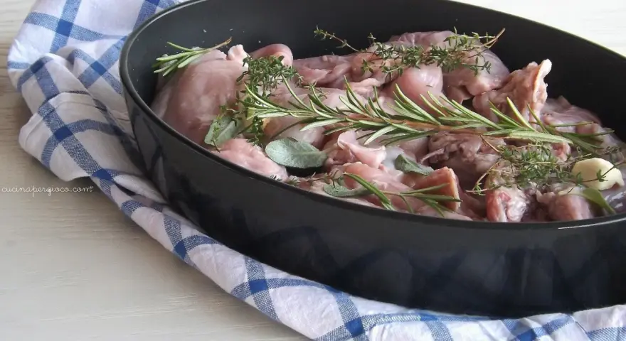 Pezzi di coniglio crudo pronti per essere cucinati al forno, profumati con rosmarino, salvia e aglio.