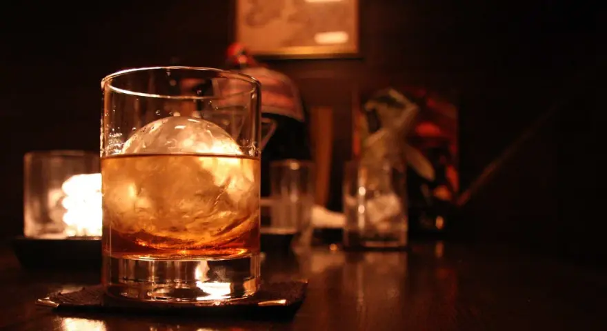 Ein Glas mit bernsteinfarbenem Whisky und einer großen Eiskugel steht auf einem dunklen Holztisch. Man trinkt Whisky mit Eis für den perfekten Genuss.
