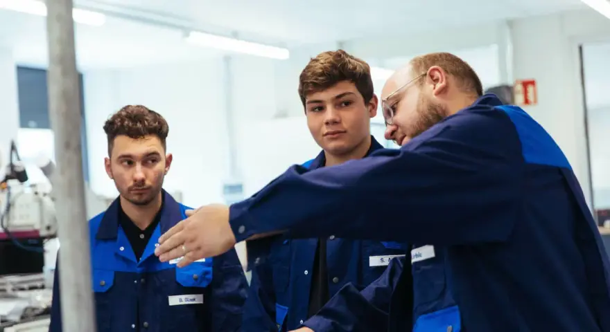 Drei junge Männer in blauen Arbeitskitteln lernen gemeinsam. Ein Ausbilder erklärt etwas, während die genz-Auszubildenden aufmerksam zuhören.
