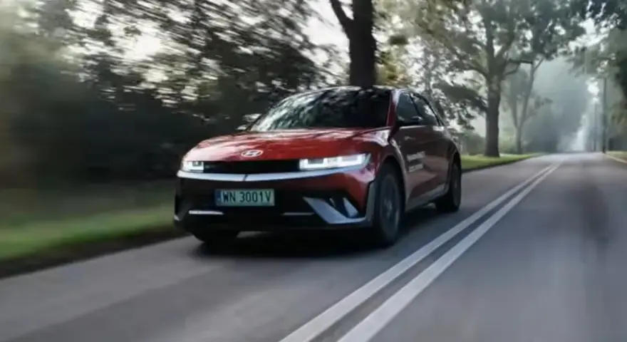 Czerwony Hyundai Ioniq 5 jedzie po wiejskiej drodze wśród drzew.