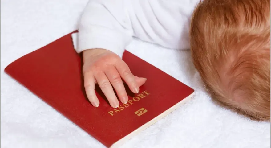 Dziecko z rudymi włosami śpi na białym ręczniku, jego dłoń spoczywa na czerwonym paszporcie. Czy do Hiszpanii potrzebny jest paszport?