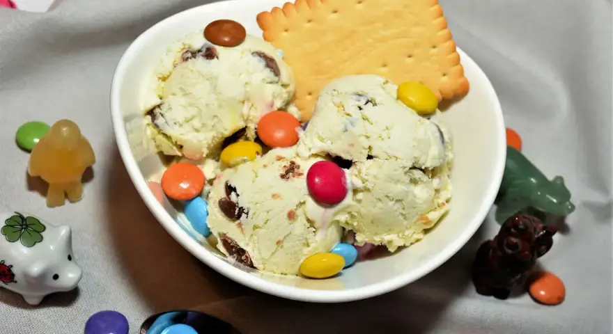 Schüssel mit cremigem Eis, garniert mit bunten Smarties und einem Keks. Kleine Figuren schmücken die Szene.