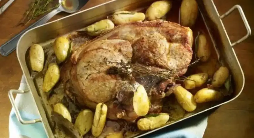 Rouelle de porc rôtie au four, entourée de pommes de terre dorées et d'herbes aromatiques. Un plat savoureux prêt à être dégusté.