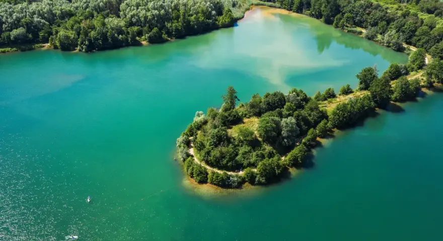 Türkisblaues Wasser umgibt eine grüne Insel mit einem Pfad am Niedernberger See. Ein Stand-Up-Paddler genießt die Ruhe.