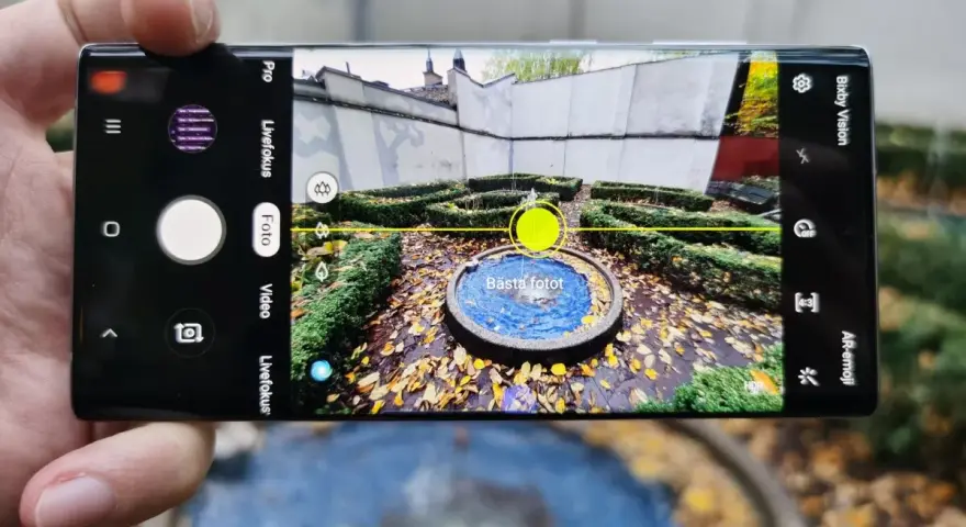 En hand håller en Samsung Galaxy Note 10 Plus med kameran öppen, redo att ta en bild av en fontän i en höstträdgård.