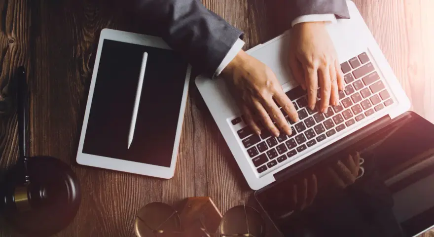 Dłonie na klawiaturze laptopa, obok tablet i młotek sędziowski. Profesjonalne narzędzia do pracy, być może związane z **taxat windykacja**.