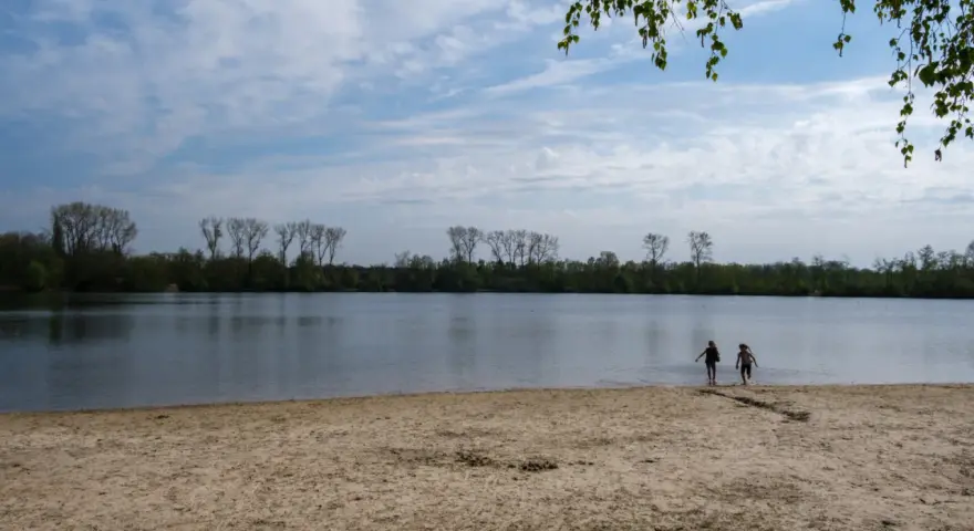 Zwei Kinder planschen im Wasser am Sandstrand des Dieksees in Lingen. Bäume säumen das Ufer unter einem bewölkten Himmel.