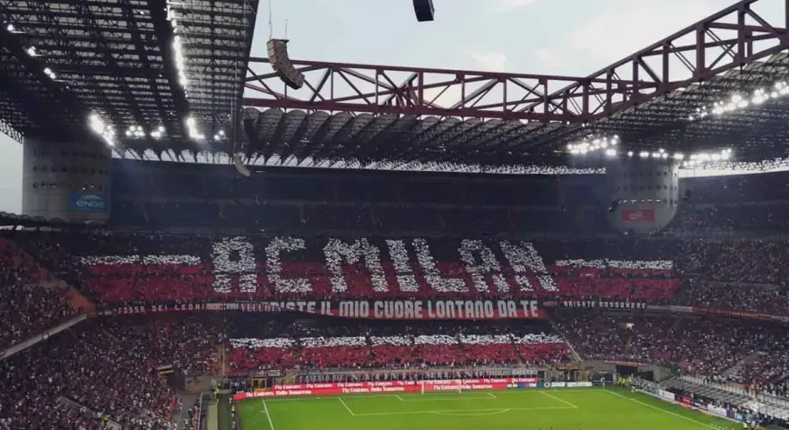 Trybuna stadionu San Siro w Mediolanie wypełniona kibicami AC Milan, tworzącymi napis "AC MILAN" i transparent "FOSSI DEI LEONI, VECCHIESTE IL MIO CUORE LONTANO DA TE".