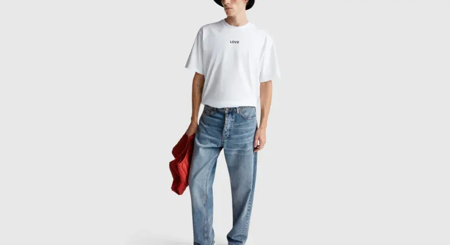 Uomo con t-shirt bianca "LOVE", cappello bucket grigio e jeans relaxed fit. Il significato di questi jeans è comfort e stile casual.
