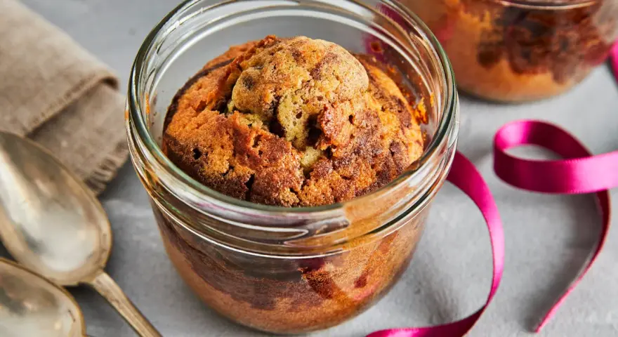 Zwei köstliche Kuchen im Glas, mit Schokoladenstückchen, warten auf Löffel. Eine pinke Schleife und Leinentuch deuten auf ein besonderes Geschenk hin.
