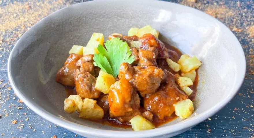 Delicioso cordero al chilindrón en un cuenco gris, acompañado de patatas fritas y una ramita de perejil.