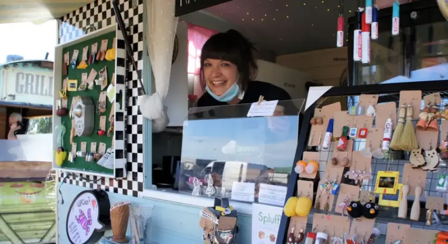 Lachende Verkäuferin auf dem Flohmarkt Friedrichshagen präsentiert handgefertigten Schmuck, darunter Eulen-Ohrringe und Miniatur-Flaschen.