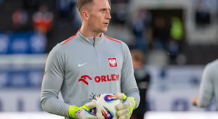 Bramkarz w szarej bluzie z orłem i napisem ORLEN, trzymający piłkę. Kolejny z polscy piłkarze za granicą, gotowy do gry.