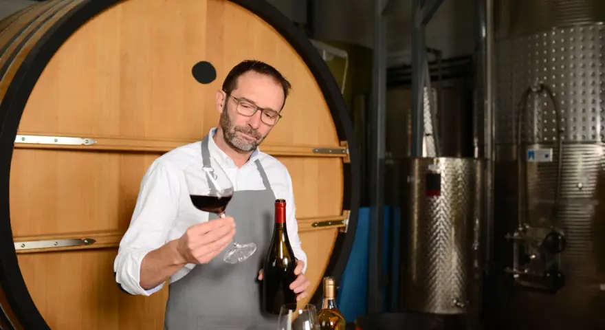 Un enologo esperto valuta un bicchiere di vino rosso, con una bottiglia in mano, davanti a una grande botte di legno e serbatoi d'acciaio.
