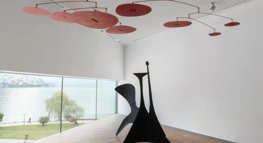 Un mobile Calder rouge flotte au-dessus d'une sculpture noire monumentale, dans une salle d'exposition lumineuse avec vue sur un lac.