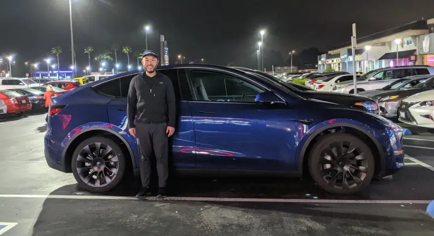 Mężczyzna stoi obok ciemnoniebieskiego Tesli Model Y na nocnym parkingu.