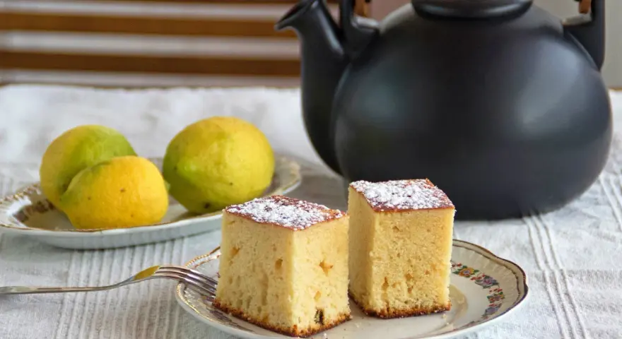 Dos porciones de bizcocho de limón espolvoreadas con azúcar glas, junto a limones frescos y una tetera negra.