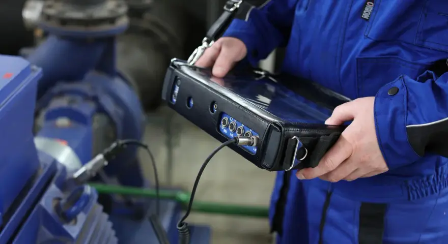 Technik w niebieskim kombinezonie wykonuje pomiary drgań maszyny przemysłowej za pomocą przenośnego urządzenia.