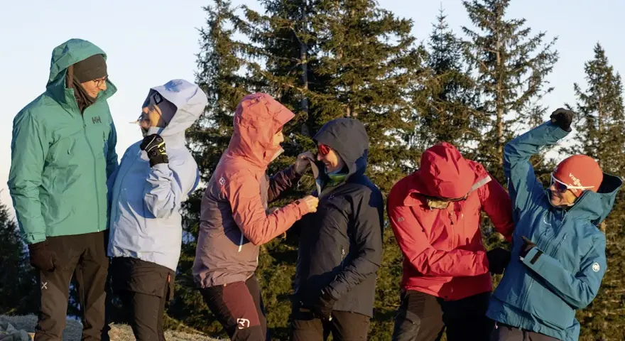 Sechs Personen in bunten Hardshelljacken stehen vor Bäumen. Eine Hardshelljacke schützt vor Wind und Wetter.