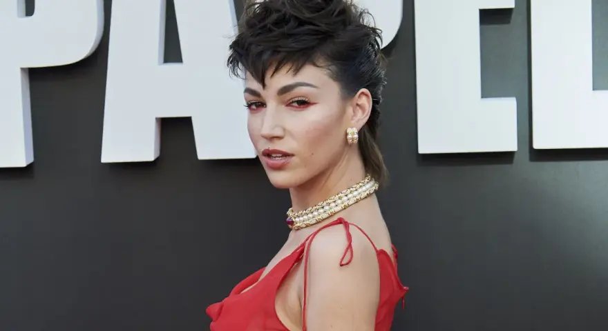 Mujer con un llamativo mullet largo, maquillaje rojo y vestido escarlata, posando ante un fondo con letras blancas.