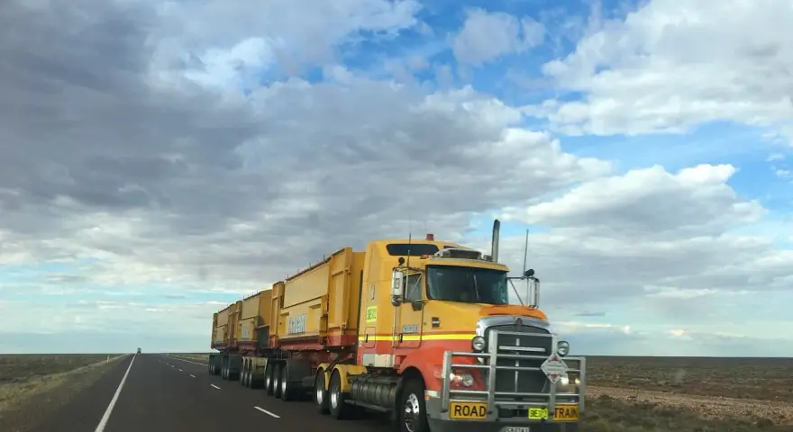 Żółta ciężarówka typu road train z naczepami na pustej drodze pod zachmurzonym niebem.