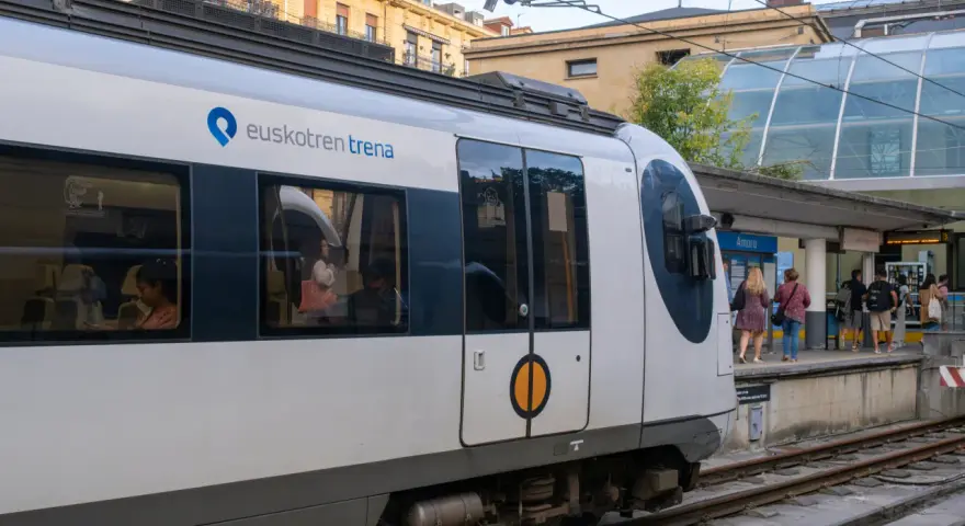 A white Euskotren train waits at the Amara station, ready to take passengers from Hendaye to San Sebastian. People stand on the platform, some boarding.