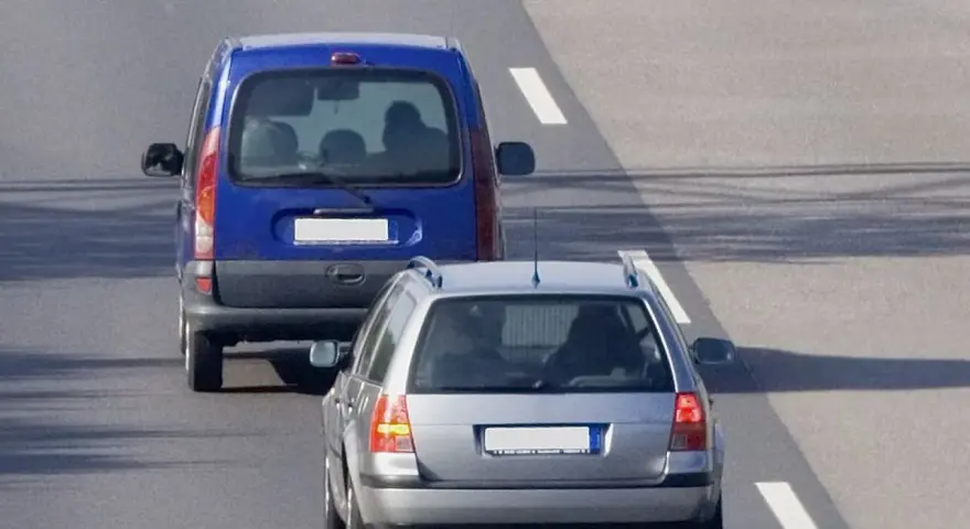 Un veicolo che precede, una monovolume blu, è seguito da un'auto station wagon argentata su una strada asfaltata con segnaletica bianca.