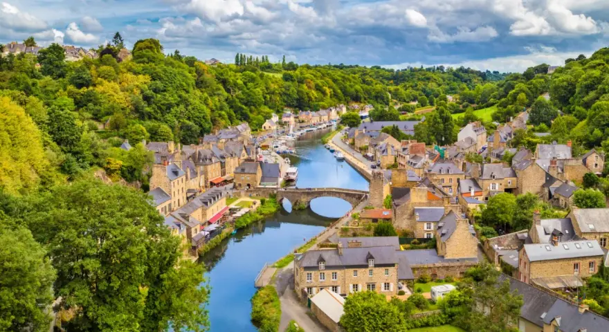 A picturesque view of Dinan, France, with its charming stone buildings lining a winding river, a stone bridge, and lush green hills under a cloudy sky.