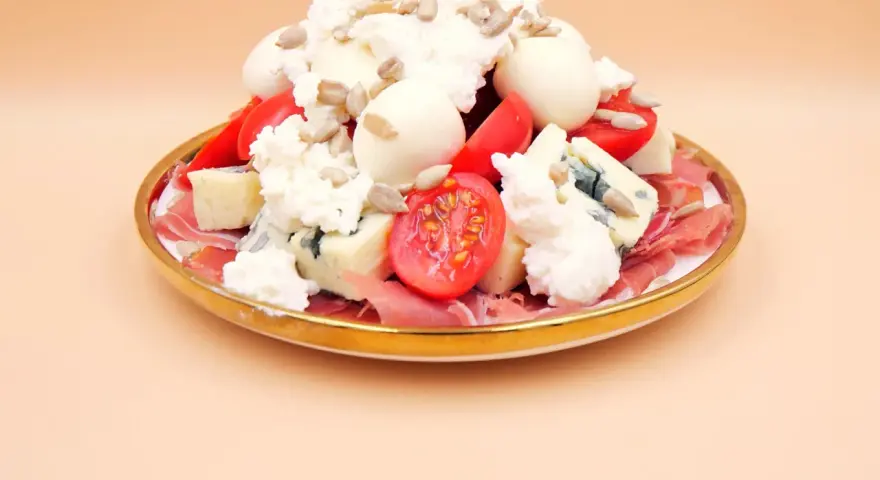 Wykwintna sałatka z szynką parmeńską, serami pleśniowymi, mozzarellą, pomidorkami i słonecznikiem na złotym talerzu.