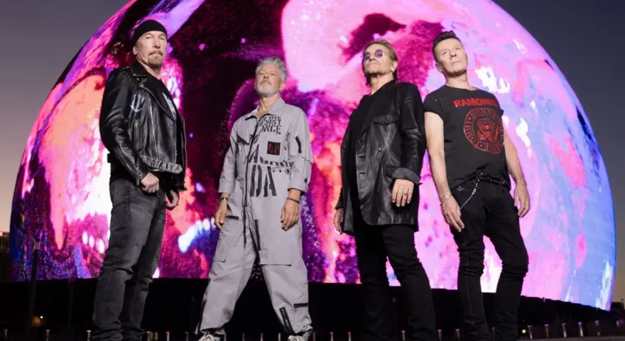 U2 w nowej płycie. Czterech członków zespołu stoi przed gigantycznym, kolorowym ekranem.