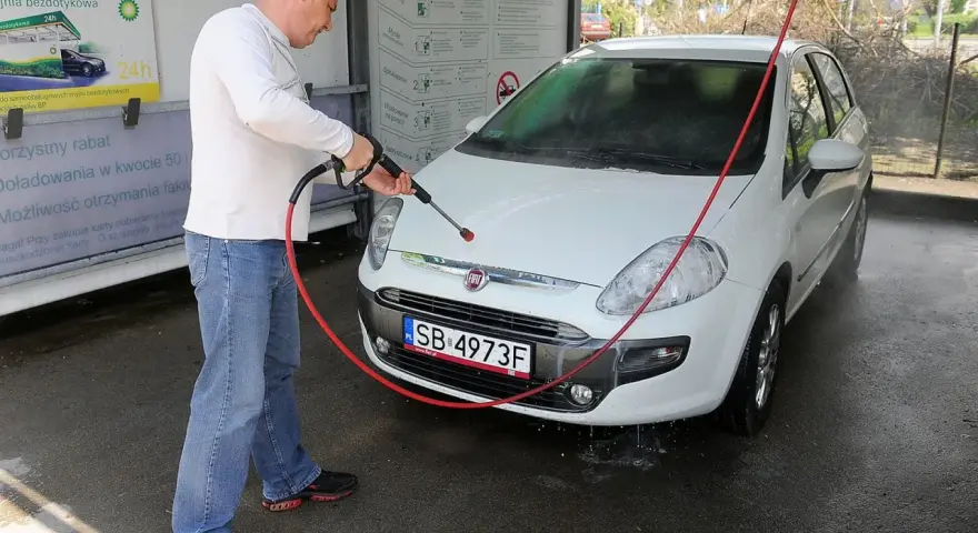 Mężczyzna myje białe auto na myjni. Na tablicy informacyjnej widnieje napis "Możliwość otrzymania faktury", sugerujący, że na myjni można płacić kartą.