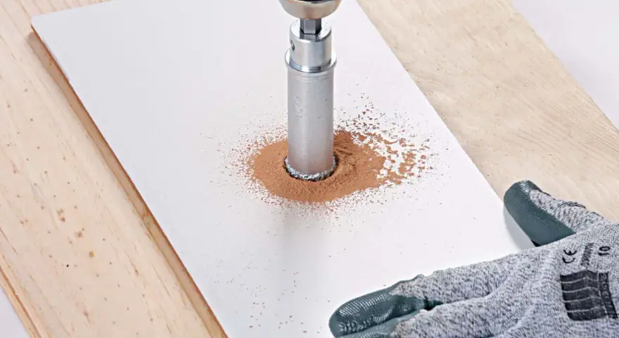 Une perceuse avec une couronne diamantée réalise une découpe carrelage, créant un nuage de poussière fine sur une plaque blanche.