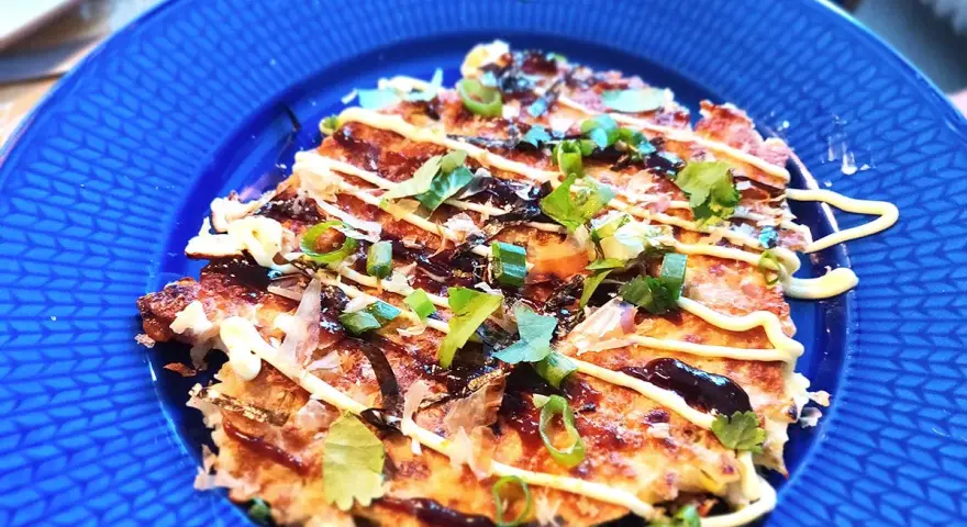 En läcker okonomiyaki på en blå tallrik, toppad med majonnäs, salladslök och torkad sjögräs. Ett perfekt okonomiyaki recept!