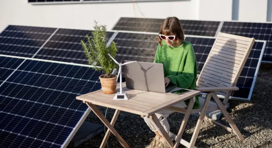 Dziewczyna w okularach pracuje na laptopie przy stole, otoczona panelami słonecznymi. Obok stoi model wiatraka, symbolizujący energię oze.
