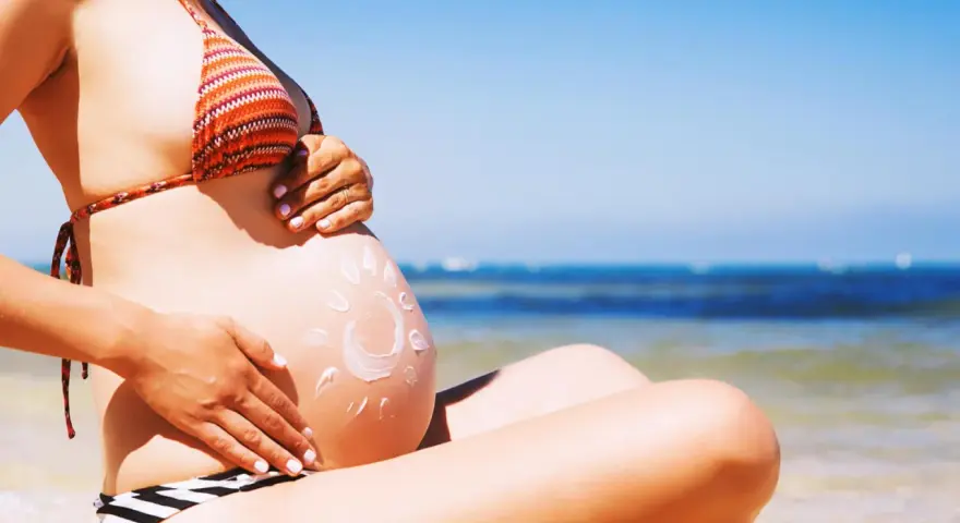 Ciężarna kobieta na plaży, z kremem przeciwsłonecznym na brzuchu w kształcie słońca. Bezpieczne opalanie natryskowe w ciąży.