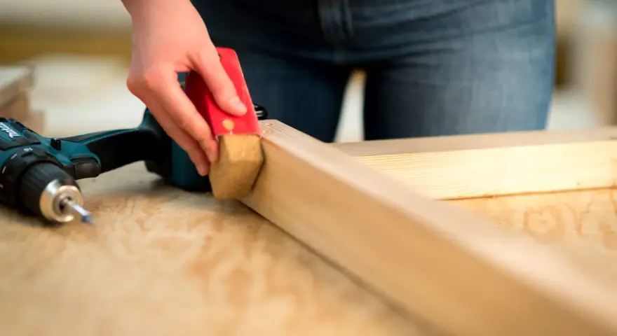 Eine Hand schleift mit einem roten Schleifklotz ein Holzstück. Daneben liegt ein Akkuschrauber.