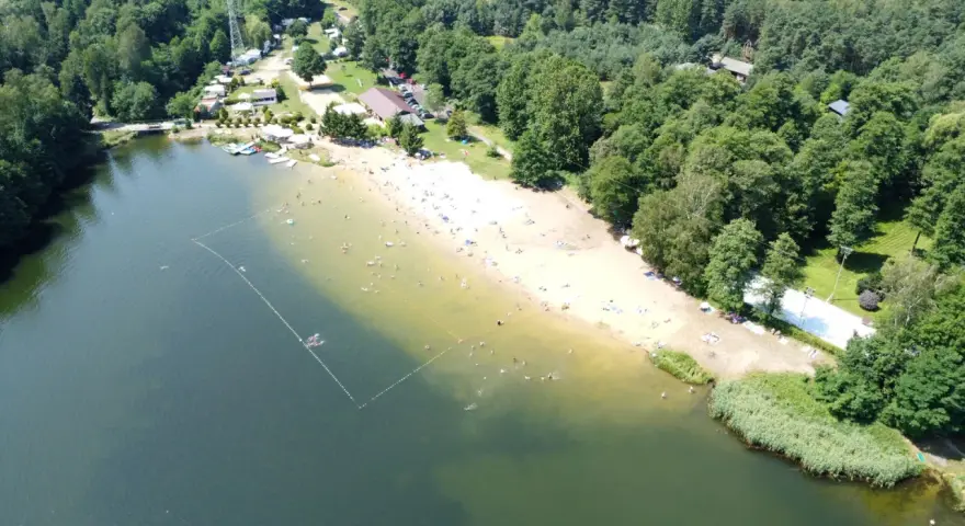 Słoneczny dzień nad jeziorem Rogoźnik. Ludzie kąpią się i opalają na piaszczystej plaży, otoczonej zielonym lasem.