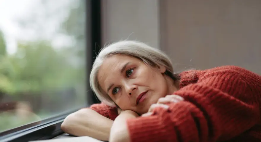 Donna con capelli grigi guarda fuori dalla finestra, pensierosa. Potrebbe cercare rimedi per la depressione estiva.