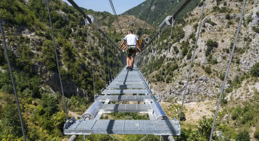 Un ponte tibetano in Puglia attraversa una gola verdeggiante, con persone che camminano su passerelle metalliche sospese tra montagne.