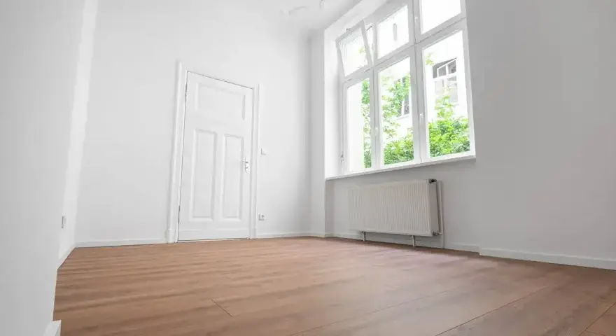 Puste, jasne pomieszczenie z białymi ścianami, drzwiami i oknem. Drewniana podłoga i grzejnik sugerują, że to gotowe do zamieszkania mieszkanie socjalne.