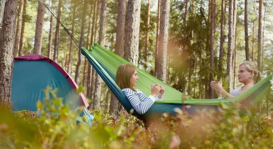 Två tjejer njuter av en lugn stund i en hängmatta på en av Sveriges fina campingar. Bredvid dem står ett tält.