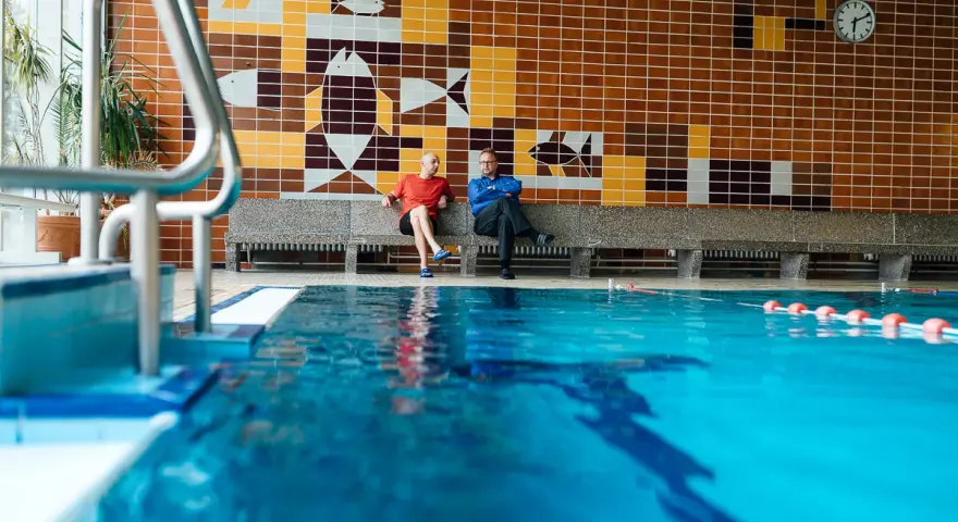 Zwei Männer sitzen in der Schwimmhalle Anklam auf einer Bank vor einer bunten Kachelwand mit Fischmotiven.